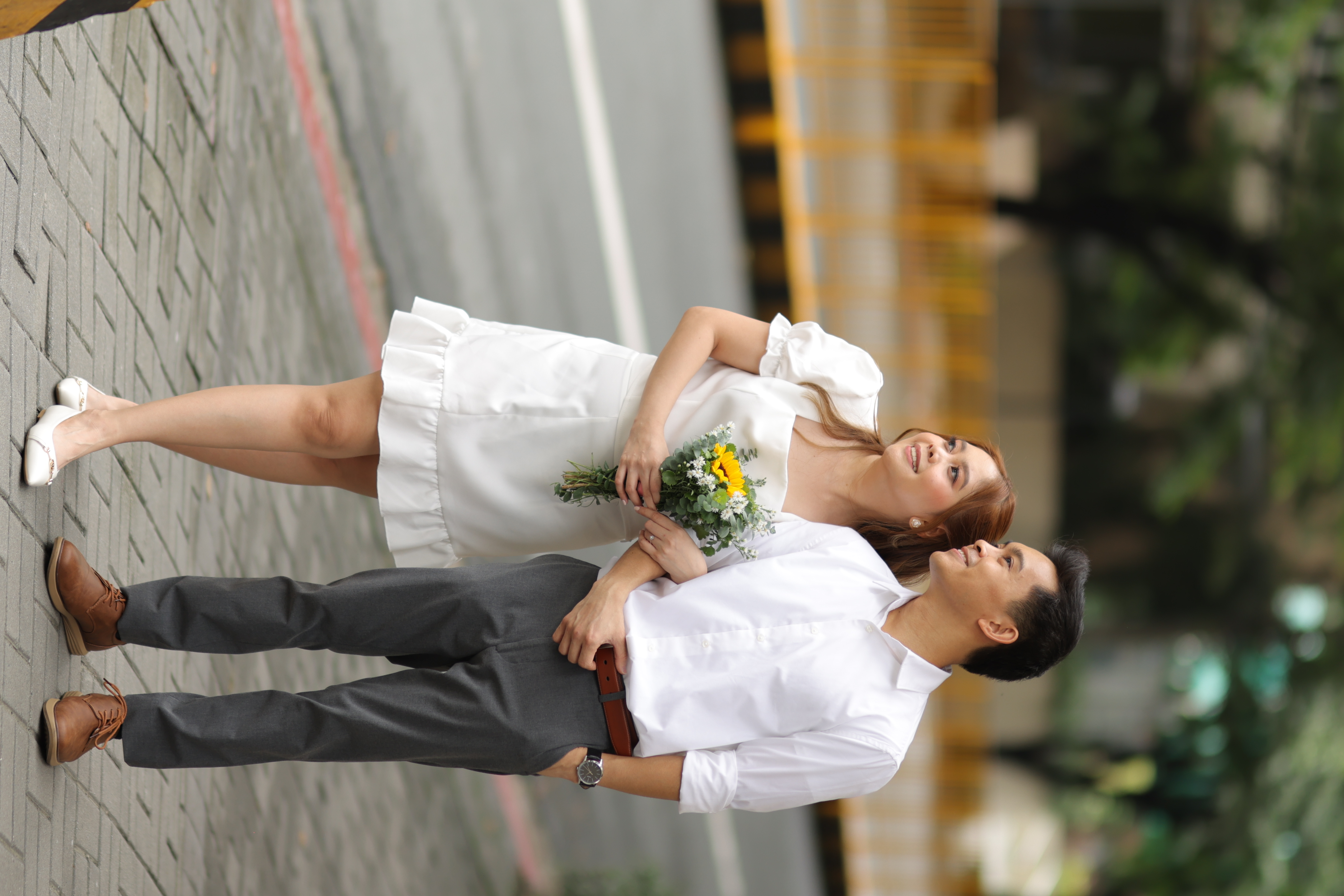 Joel and Irianne standing arm in arm on a brick plaza, Irianne holding a sunflower bouquet
